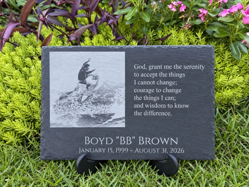 Personalized black slate memorial plaque with an engraved surfing photograph, name, dates and prayer, displayed on a stand in a garden.