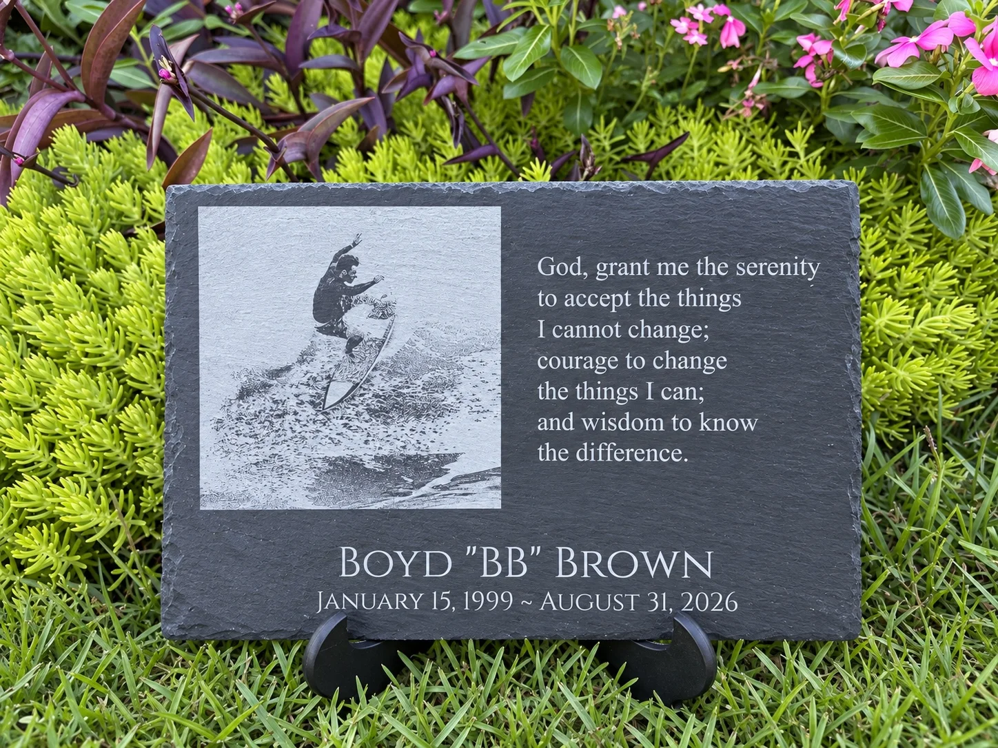 Personalized black slate memorial plaque with an engraved surfing photograph, name, dates and prayer, displayed on a stand in a garden.